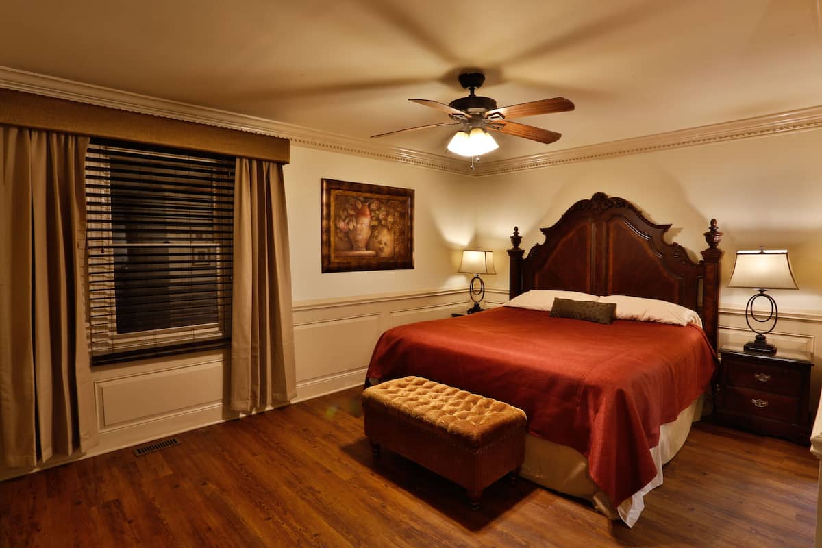 A bedroom with a wooden floor, a bed with a red bedspread, a ceiling fan, two bedside lamps, and a window with blinds.