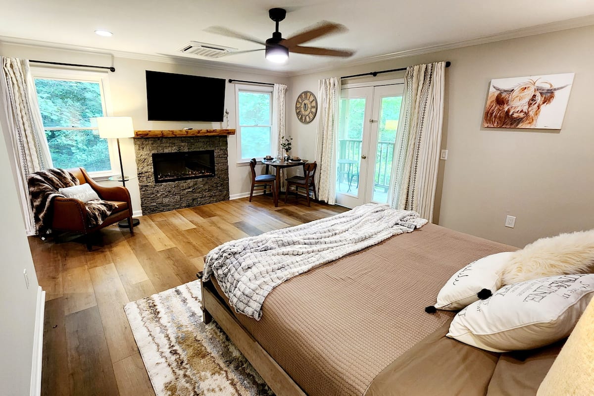 A bedroom with a large bed, a fireplace, a TV mounted on the wall, and a dining area with a table and chairs.