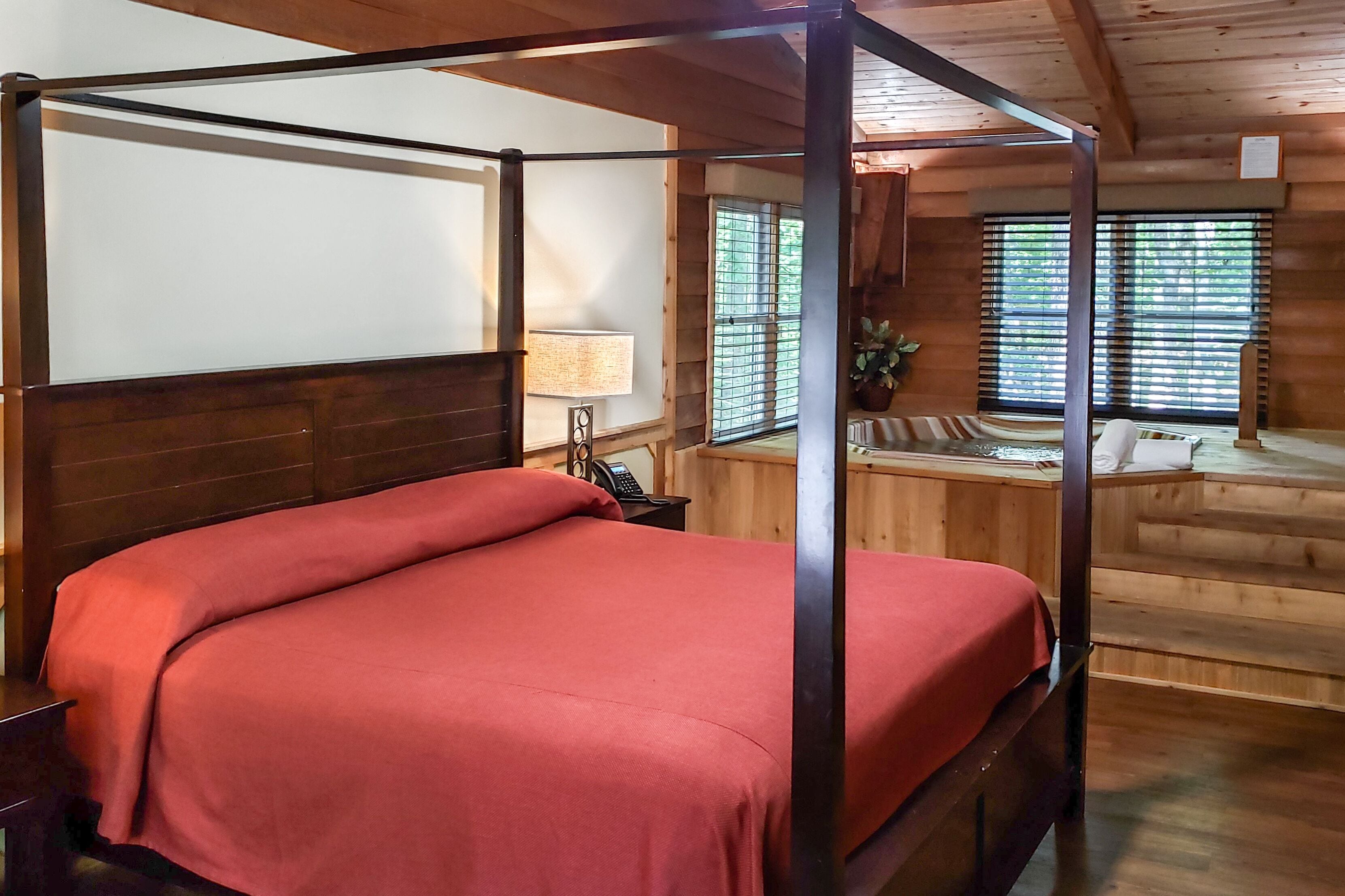 A four-poster bed with a red bedspread, a wooden headboard, and a nightstand with a lamp.