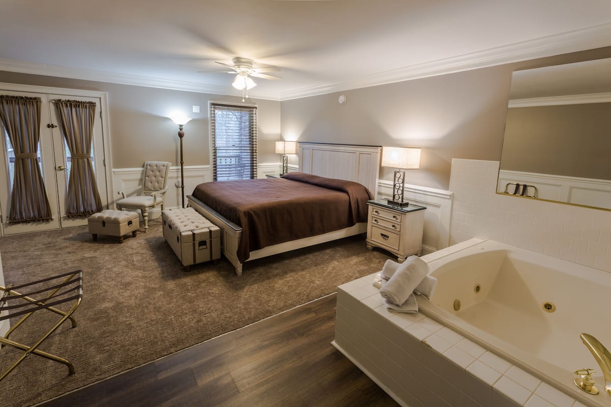 A hotel room with a large bed, a sitting area with a chair and ottoman, a desk with a lamp, a large window with curtains, a ceiling fan, and a bathtub.