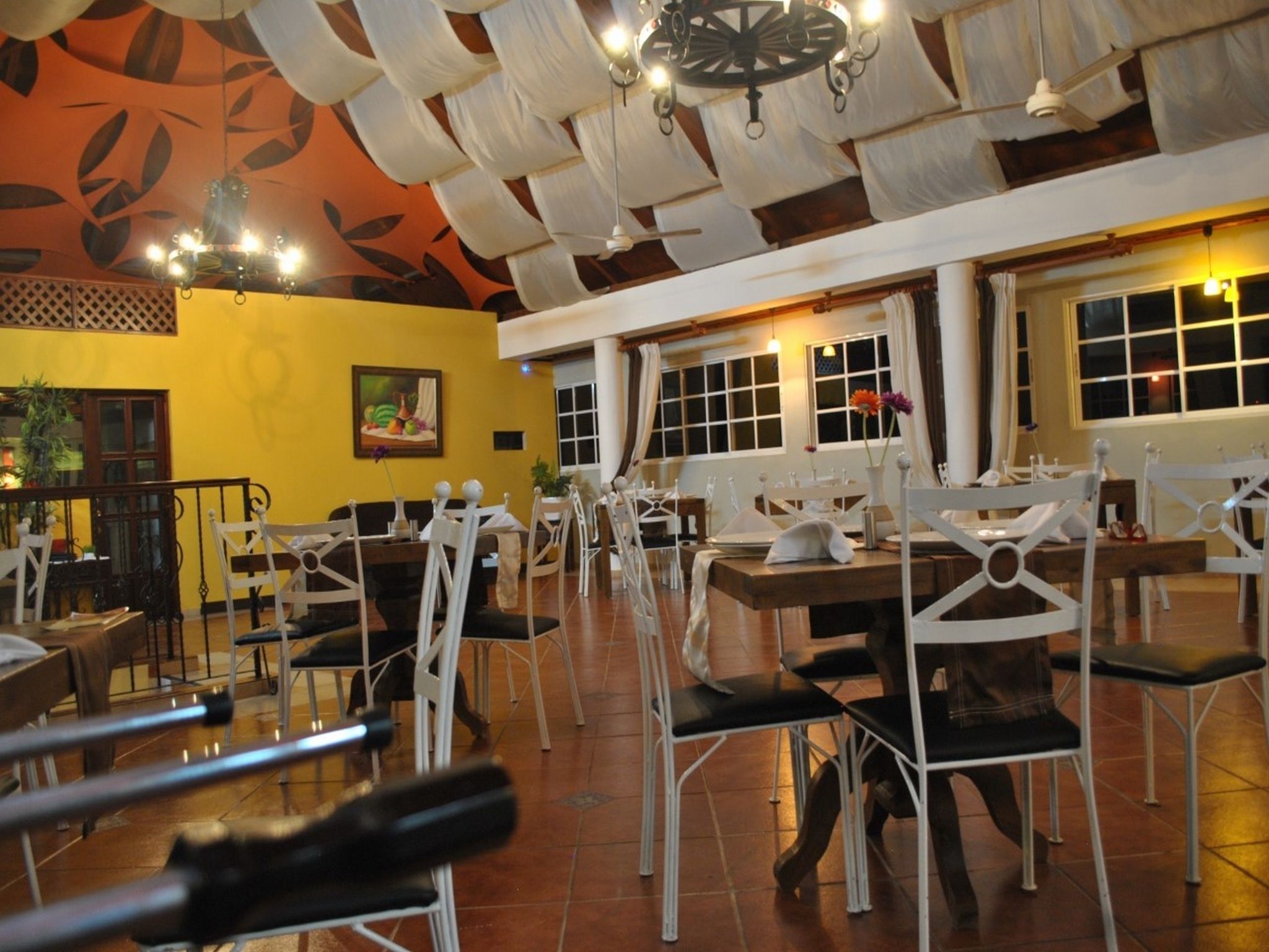 Un restaurant avec des chaises et des tables blanches, un mur jaune et un plafond orné d’un motif de feuilles.
