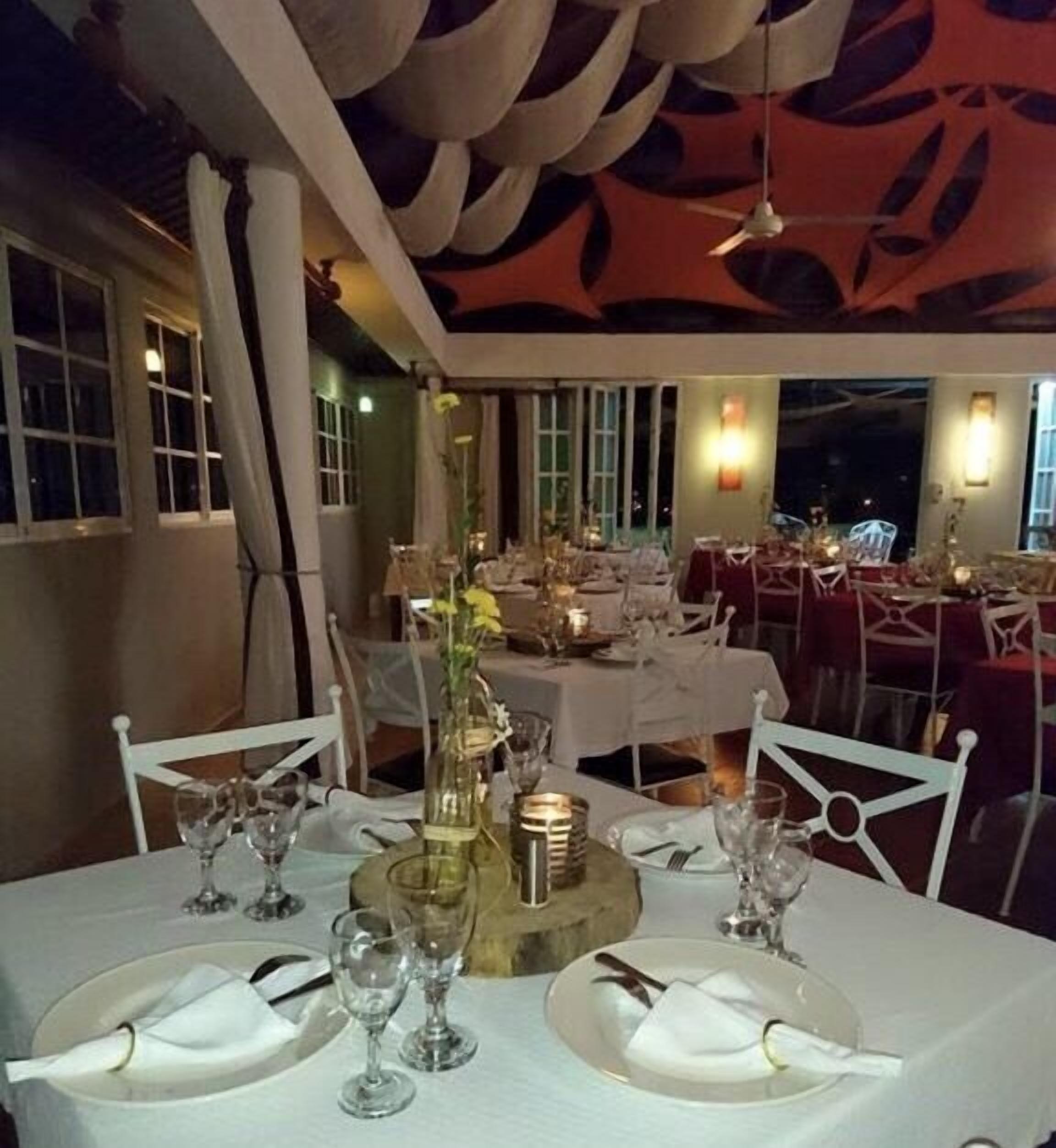 Un comedor con mesas dispuestas para una comida, con manteles blancos, copas de vino y un centro de mesa con un jarrón y flores.