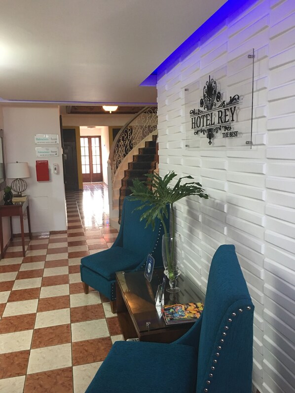 Un vestíbul d'hotel amb un paviment a quadradets, una escala amb barana ornamentada i una taula de recepció amb una planta.