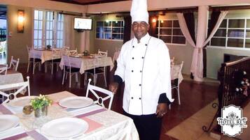 A chef in a white uniform standing in a dining area with tables set for a meal.