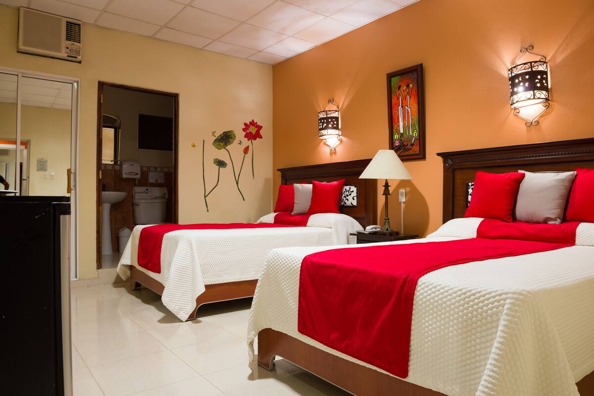 Une chambre d’hôtel avec deux lits, chacun recouvert d’une couvre-lit rouge et blanche, une table de chevet avec une lampe et un luminaire fixé au mur.