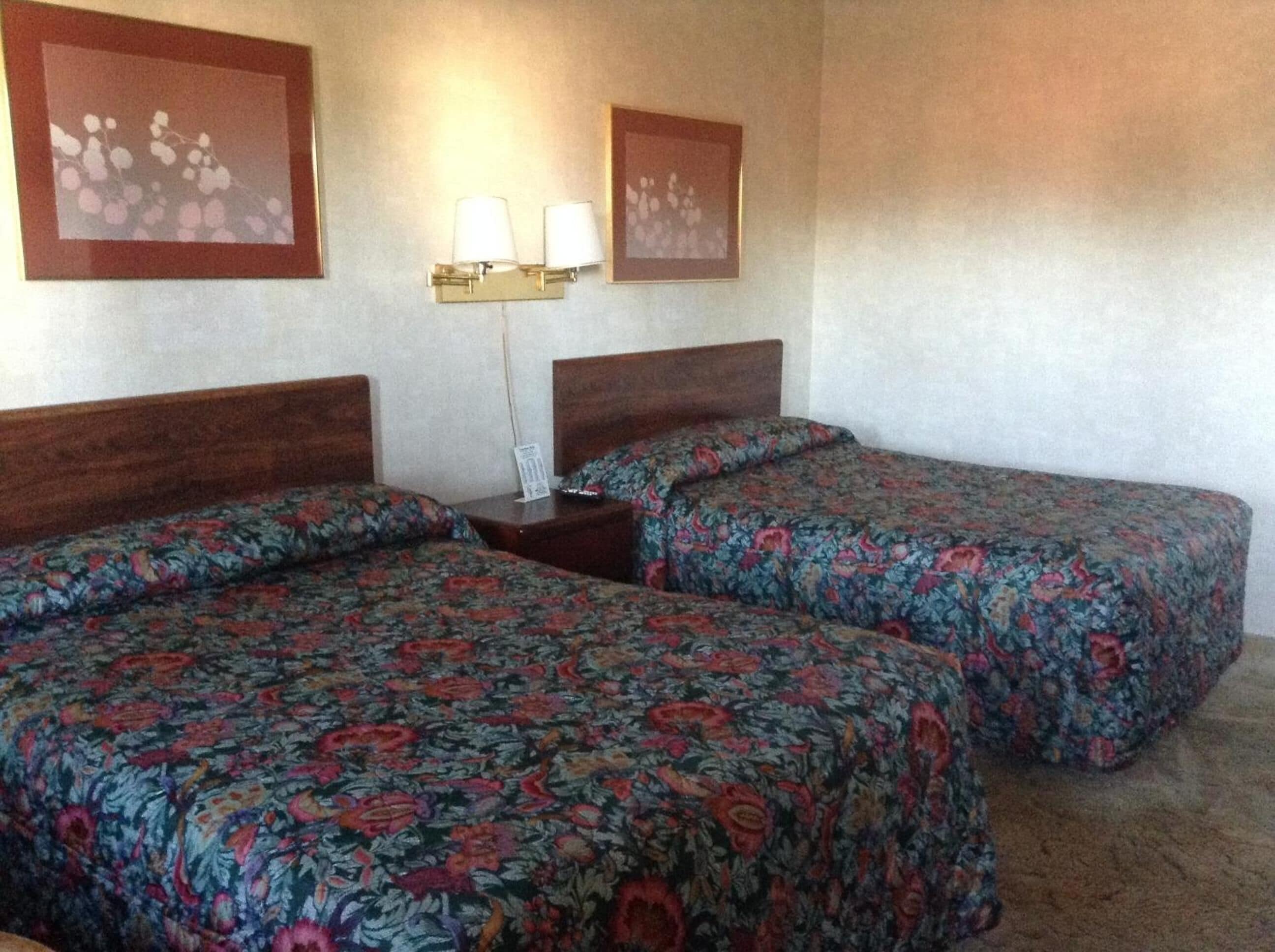 A hotel room with two single beds, floral bedspreads, a nightstand with a lamp, and two wall-mounted lamps.