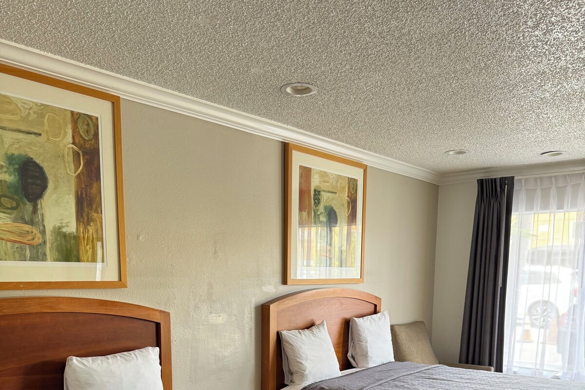 A hotel room with two beds, a wooden headboard, a nightstand, a painting, and a window with curtains.