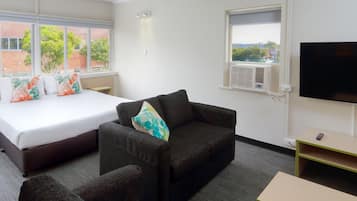 A hotel room with a bed, a sofa, a small table, a TV, and a window with an air conditioning unit.