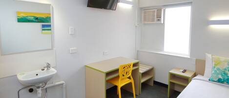 Habitación de hotel con cama, escritorio con silla, televisor montado en la pared, ventana con persianas y lavamanos con espejo.