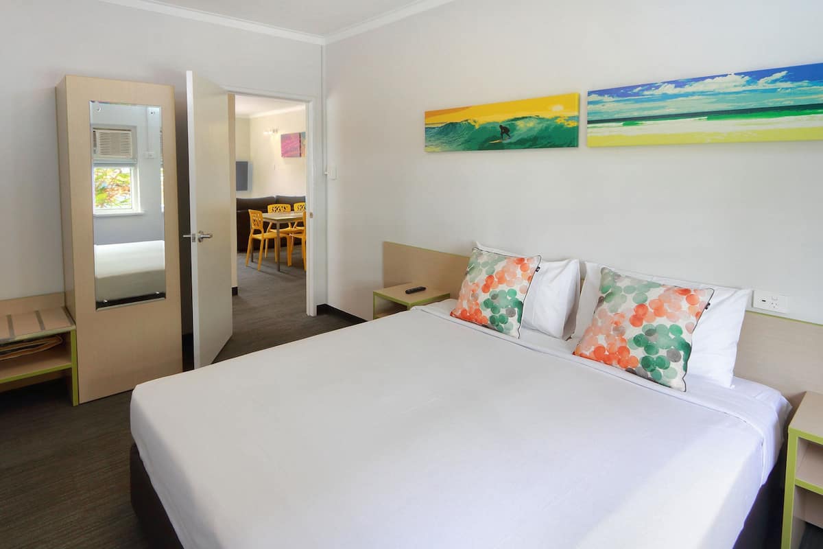 Habitación de hotel con cama, escritorio, silla y una mesita. Se ve una puerta que da a un baño y un pasillo.