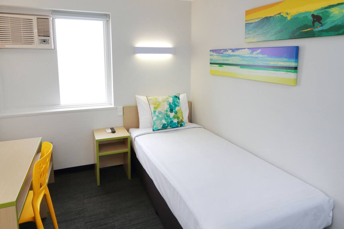 Una cama individual con colchón blanco y almohada de colores, una pequeña mesita de noche de madera, un escritorio con silla y una ventana con equipo de aire acondicionado.