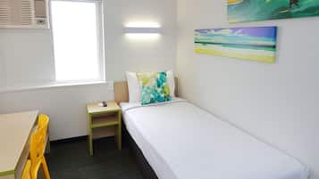 Un letto singolo con un materasso bianco e un cuscino colorato, un comodino in legno, una scrivania con una sedia e una finestra con un condizionatore d'aria.