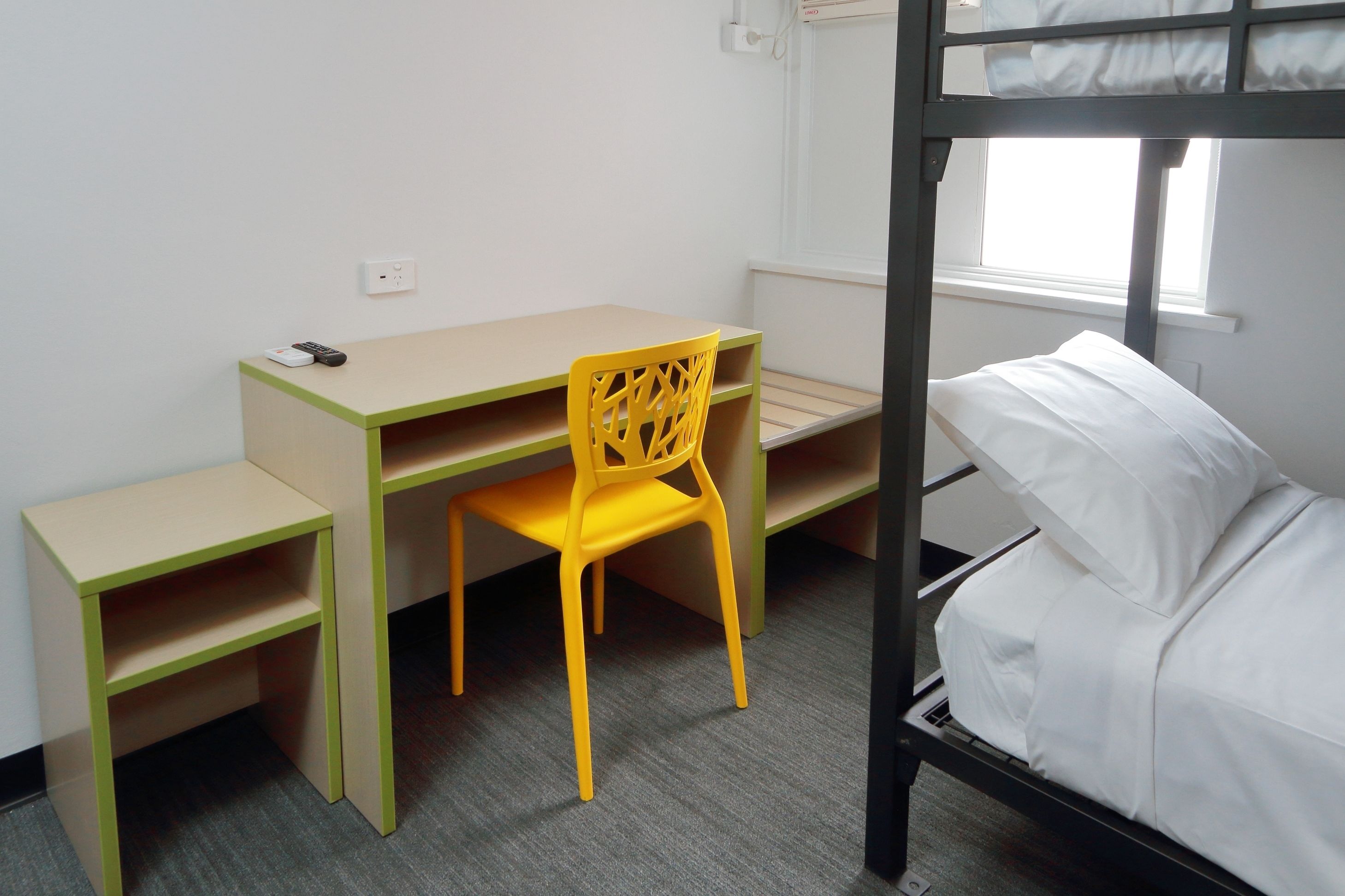 A bunk bed with a white mattress, a yellow chair, a small desk with a remote control, and a window with daylight.