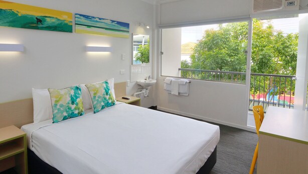 A hotel room with a bed, bedside table, a small desk with a chair, a window with a view of trees, and artwork on the wall. - Palm Beach Hotel (Palm Beach)