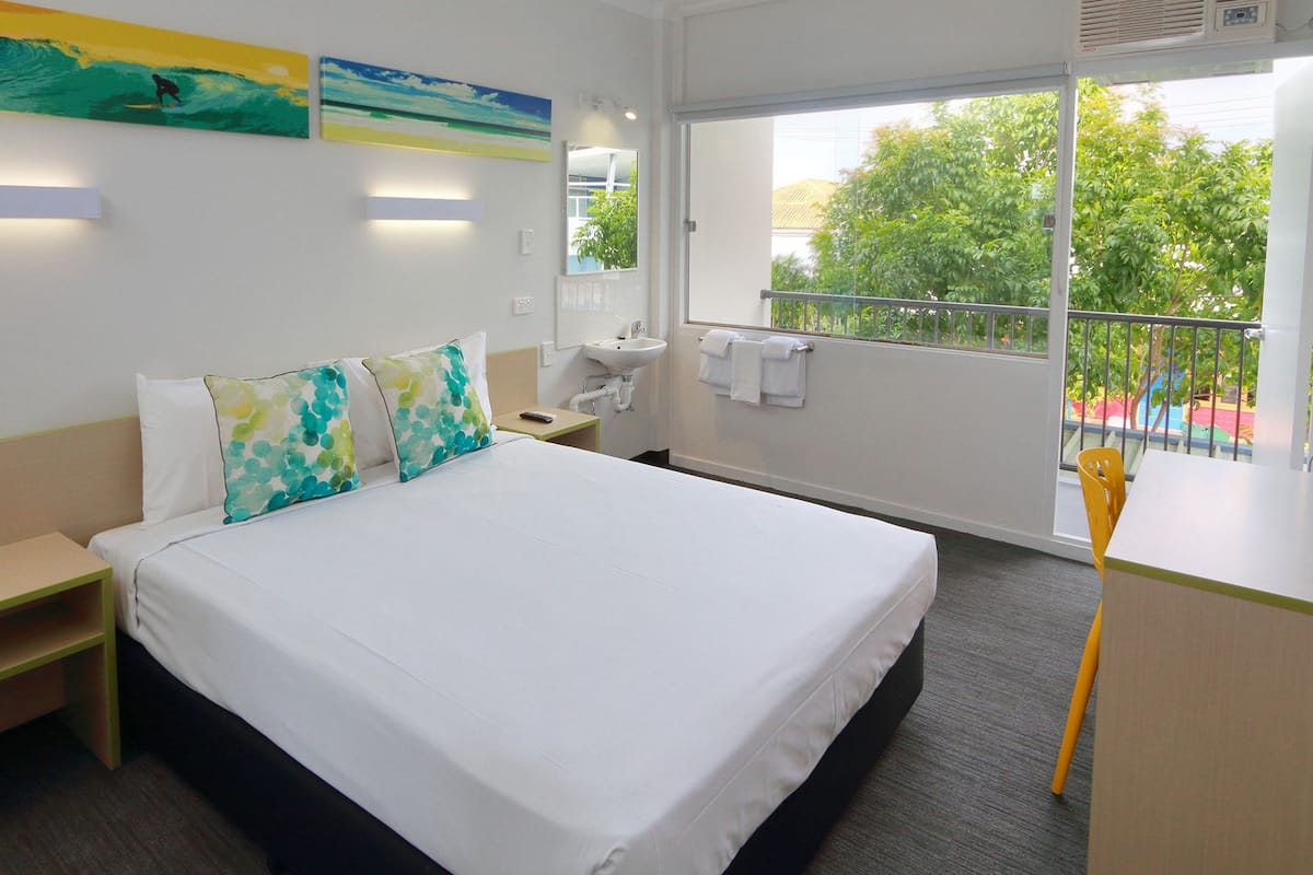 Habitación de hotel con cama, mesita de noche, un escritorio pequeño con una silla, ventana con vista a árboles y una obra de arte en la pared.