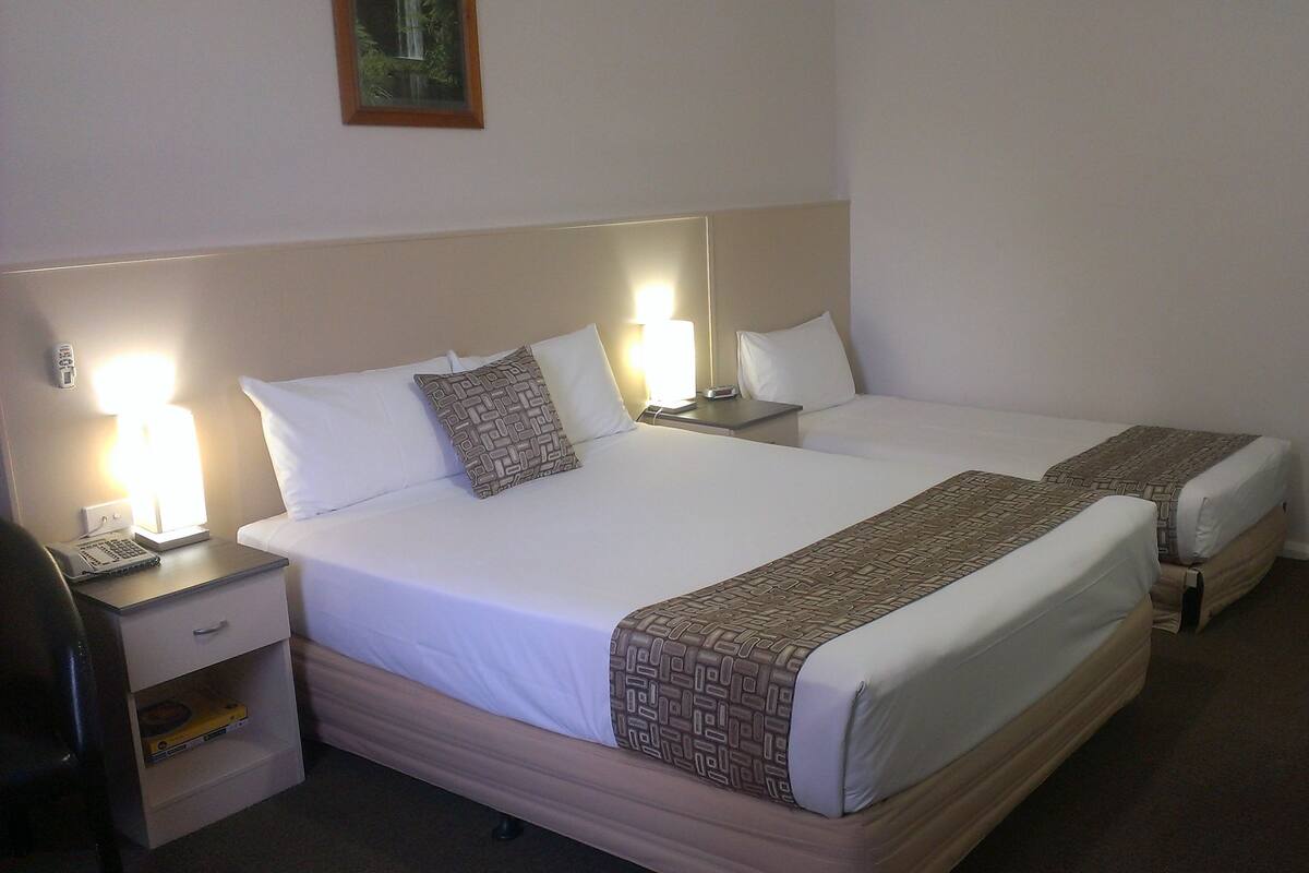 A hotel room with two beds, each with a headboard, bedside tables, and lamps. There is a chair and a desk with a lamp.