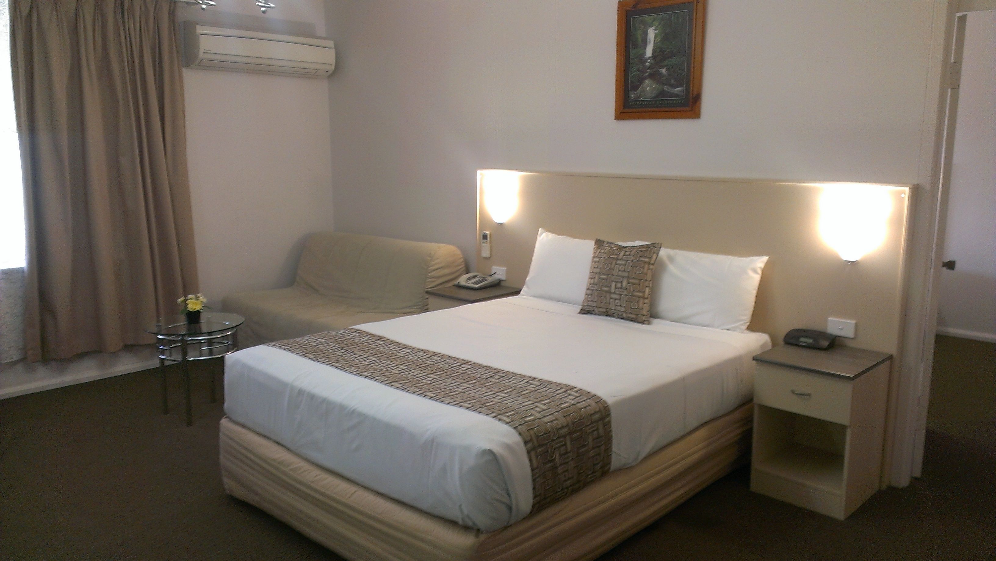 A hotel room with a large bed, a bedside table, a chair, a small table with a vase, and a wall-mounted air conditioner.