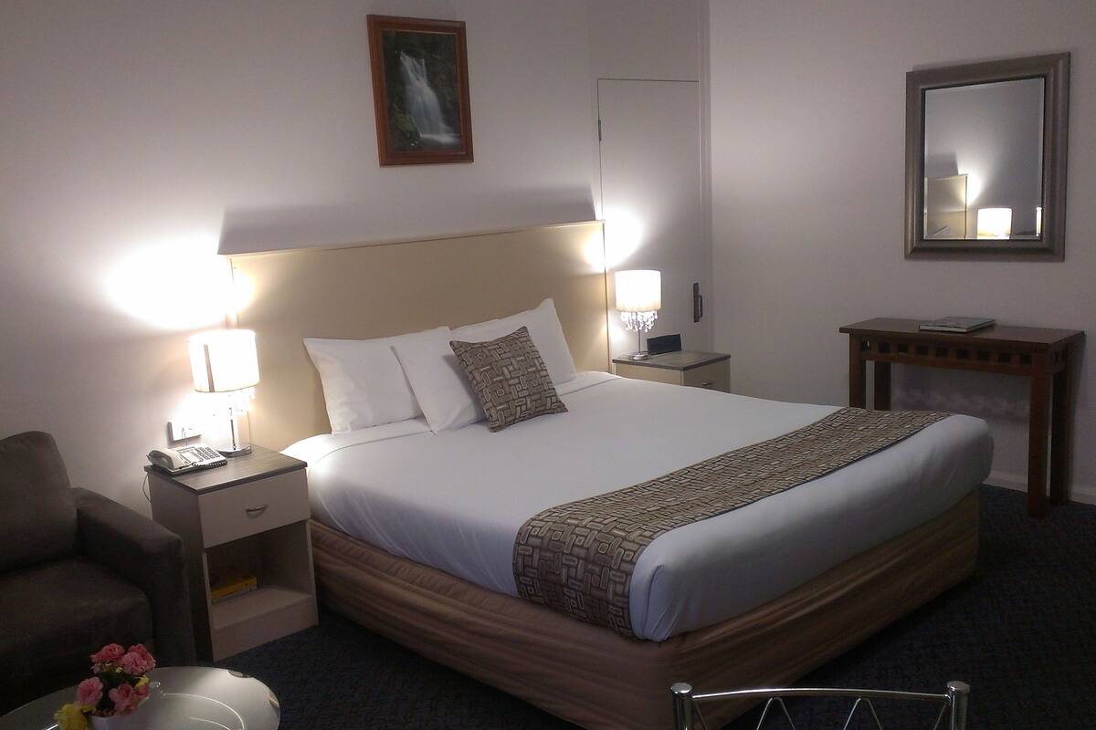 A neatly made hotel room with a large bed, two bedside lamps, a small table, a chair, and a mirror.