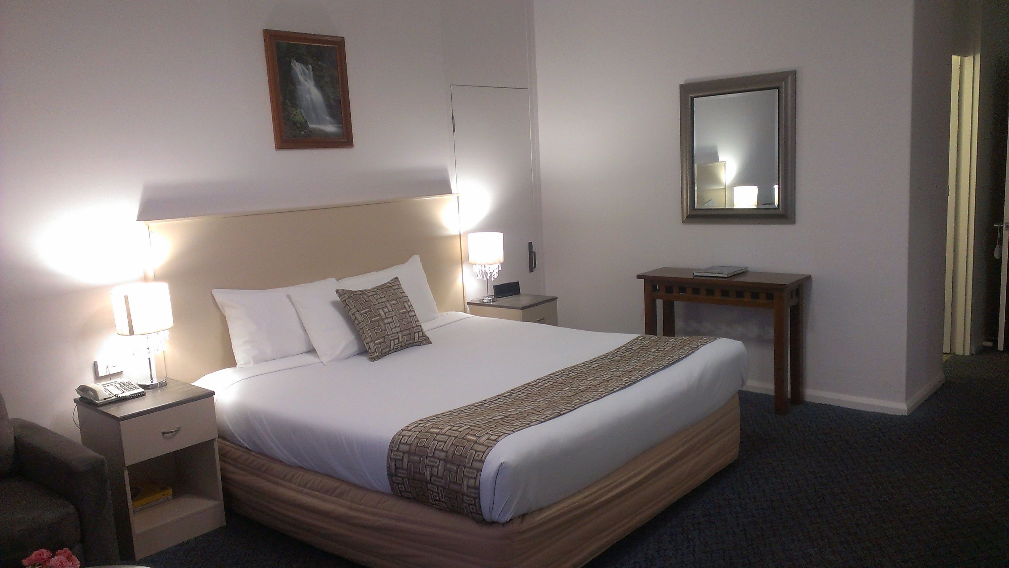 A neatly made bed with white linens and a patterned throw, two bedside lamps, a small bedside table with a phone, a nightstand, a wooden side table with a book, a mirror, and a framed picture on the wall.