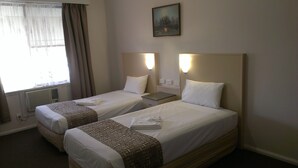 Une chambre d’hôtel avec deux lits simples, chacun étant équipé d’un oreiller et d’une couverture. Il y a une fenêtre avec des rideaux, un tableau au mur et une table de chevet avec une lampe.