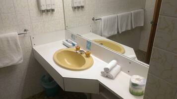 Une salle de bain avec un lavabo jaune, un comptoir blanc et un miroir. On y trouve des articles de toilette sur le comptoir, une poubelle et un porte-serviettes avec des serviettes.