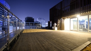 Una terraza en la azotea con cubierta de madera, barandillas de vidrio y un edificio moderno con amplios ventanales.