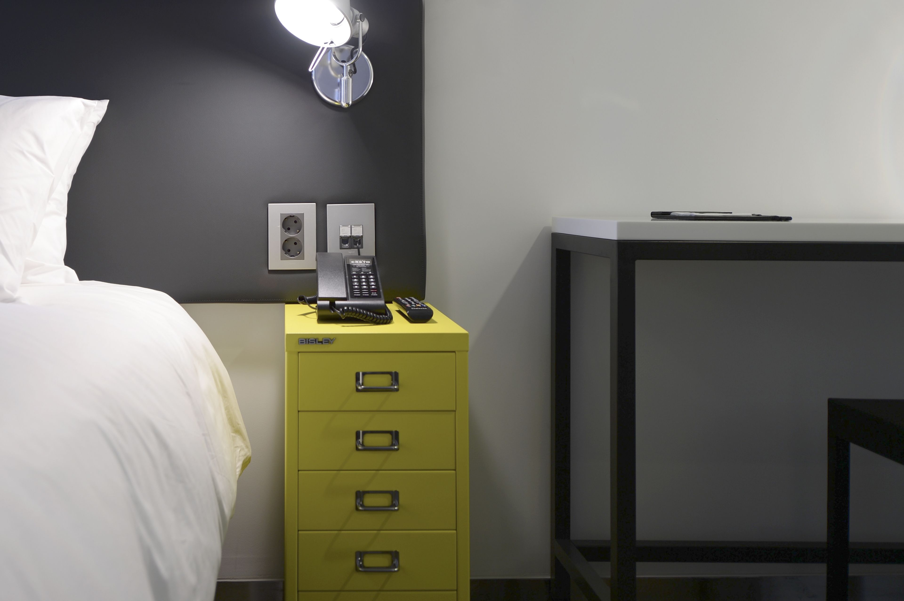 Une chambre d’hôtel avec un lit, un mini-bar jaune, un bureau avec une lampe et un téléphone posé sur le mini-bar.