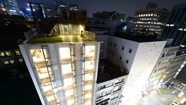 Un edificio de gran altura con varios pisos, ventanas amplias y una terraza en la azotea con barandilla.