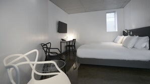 Una habitación de hotel moderna con una cama grande, un escritorio con silla, un televisor montado en la pared y una ventana que proporciona luz natural.