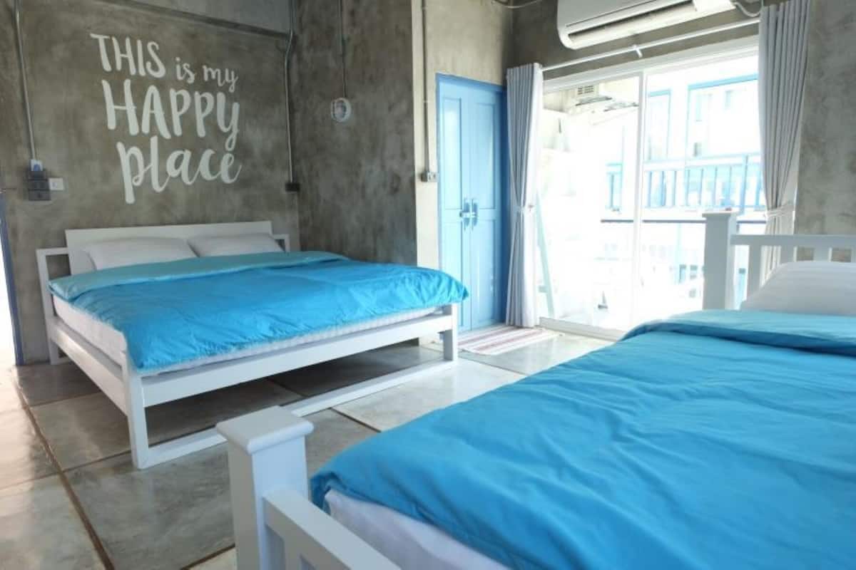 A bedroom with two beds, a concrete wall with a motivational quote, a blue door, and a balcony with a view.