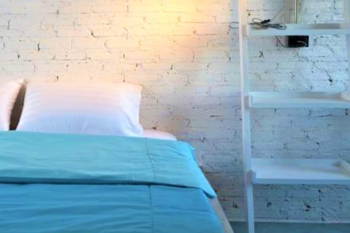 A bed with a blue bedspread and white pillows against a white brick wall.
