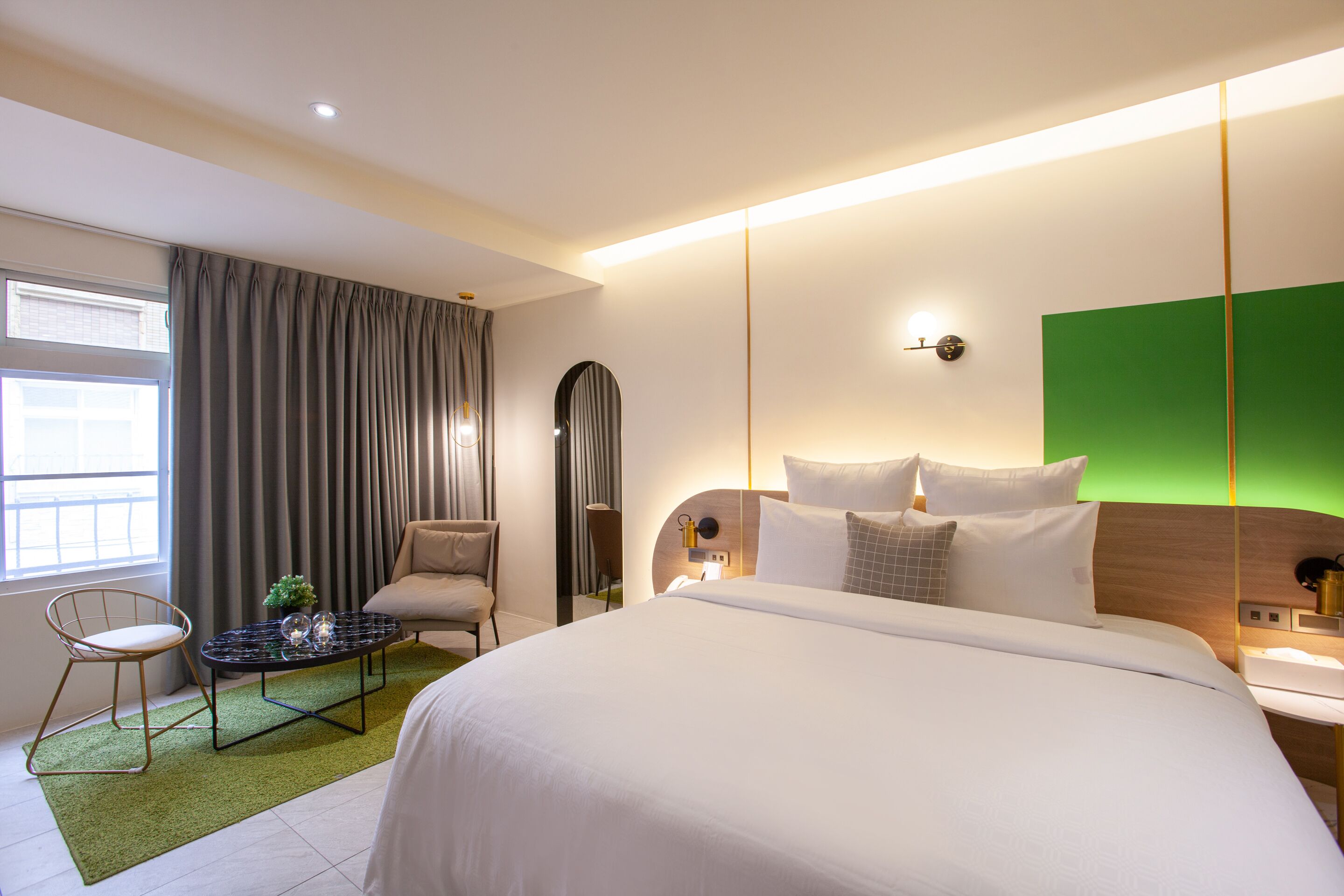 A modern hotel room with a large bed, a small round table, a chair, and a green accent wall.