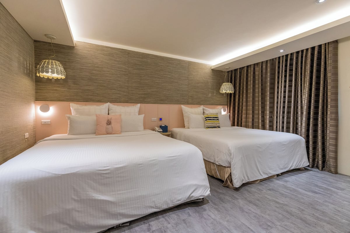Une chambre d’hôtel avec deux lits, une tête de lit en bois, une table de chevet et une grande fenêtre avec des rideaux.