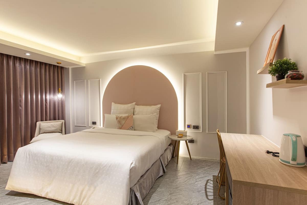 Une chambre d’hôtel moderne avec un grand lit, un bureau et une chaise. On y trouve une petite table avec une horloge et une plante accrochée au mur.