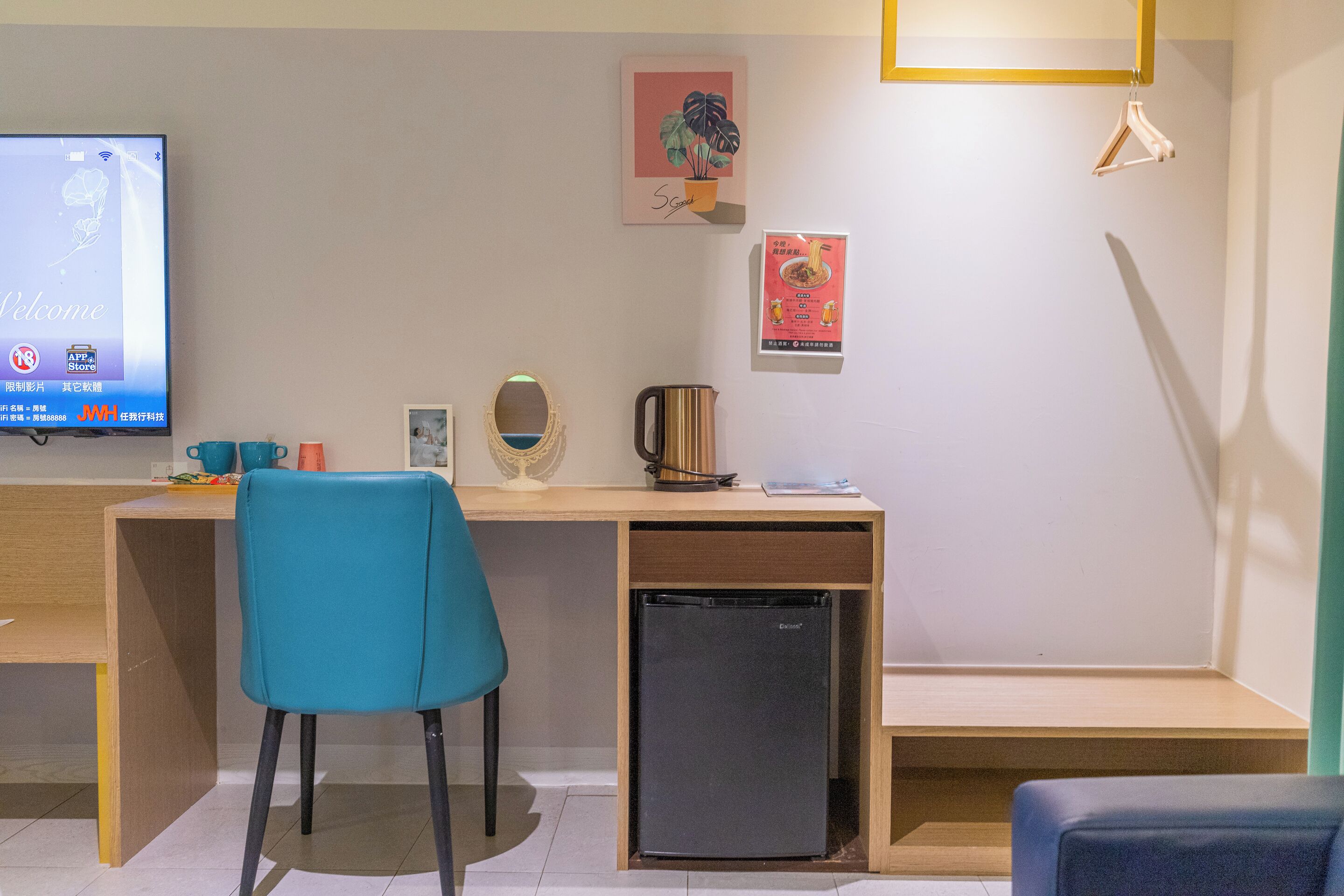 A modern room with a wooden desk, a blue chair, a small black refrigerator, a television, and wall decorations including a plant and a poster.