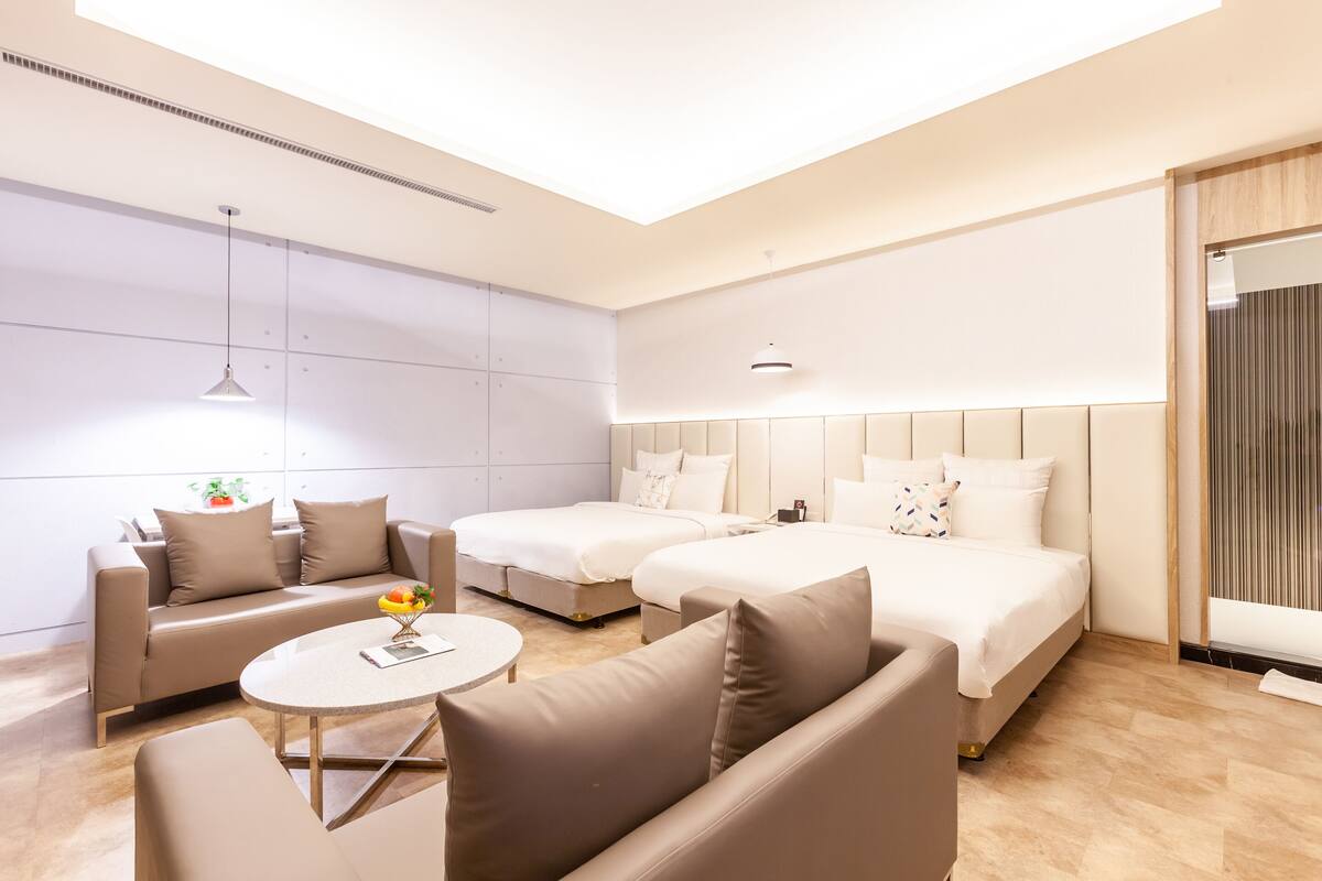 A modern hotel room with a sofa, two beds, a round table, and a large window.