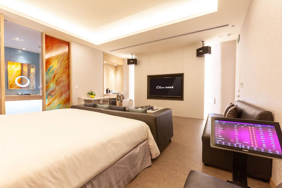 A modern hotel room with a large bed, a sofa, a desk, and a flat-screen TV.