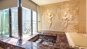 Un spa moderno con una amplia pared de piedra con esculturas en relieve, una bañera de mármol y una zona de estar con paredes de cristal y muebles de madera.