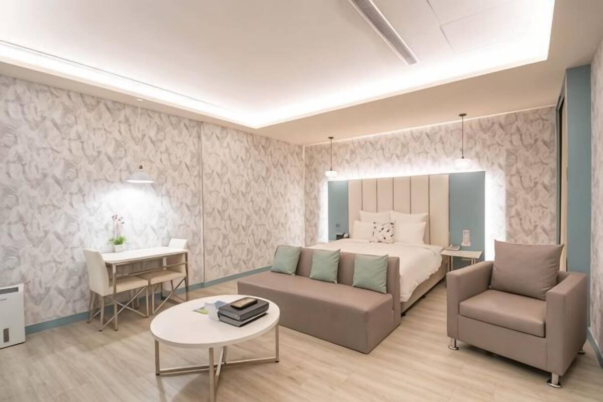 A modern hotel room with a bed, sofa, armchair, desk, and a round coffee table.