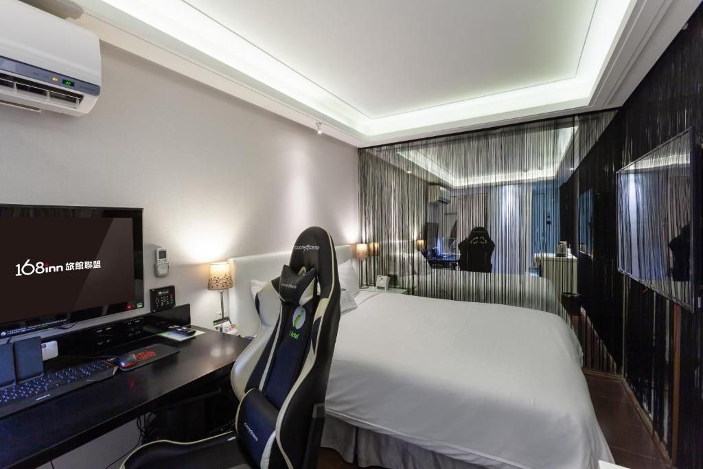 A hotel room with a bed, a desk with a computer and keyboard, a gaming chair, a TV, and a mirror reflecting the room.