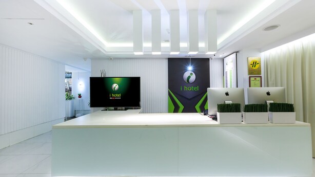 Un espace d’accueil d’hôtel au design moderne, comprenant un comptoir d’accueil, un téléviseur à écran plat et un logo sur le mur.