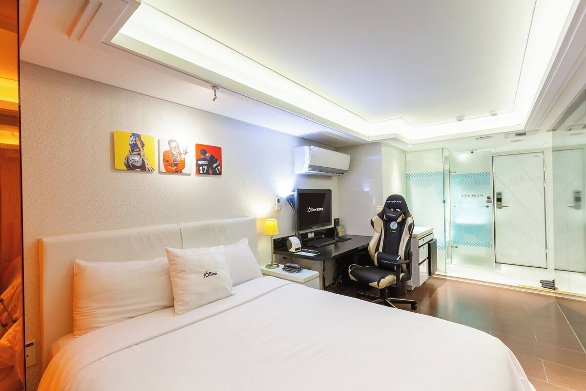Habitación de hotel con cama, escritorio, silla, televisor y puerta de vidrio que da a un baño.
