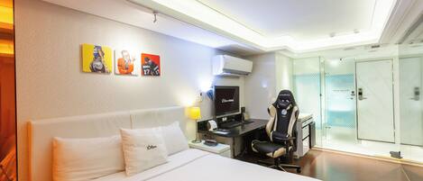 A hotel room with a bed, a desk, a chair, a TV, and a glass door leading to a bathroom.