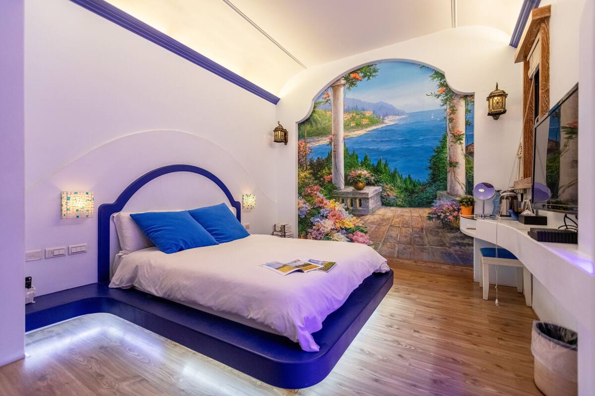 A bedroom with a bed, a desk, and a view of the sea through an archway.