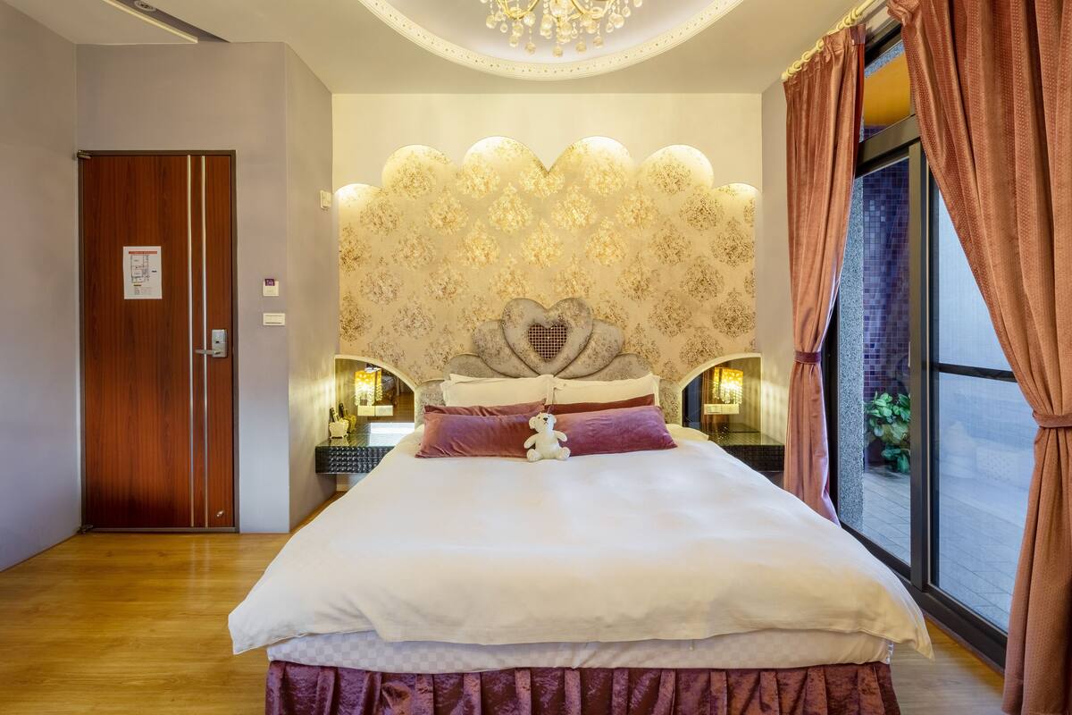 A spacious bedroom with a large bed, a decorative headboard, bedside lamps, and a view through a window with curtains.