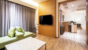 Una camera d'albergo moderna con televisione a schermo piatto montata su un pannello di legno, un divano verde con cuscini, un piccolo tavolo e un pavimento in legno.