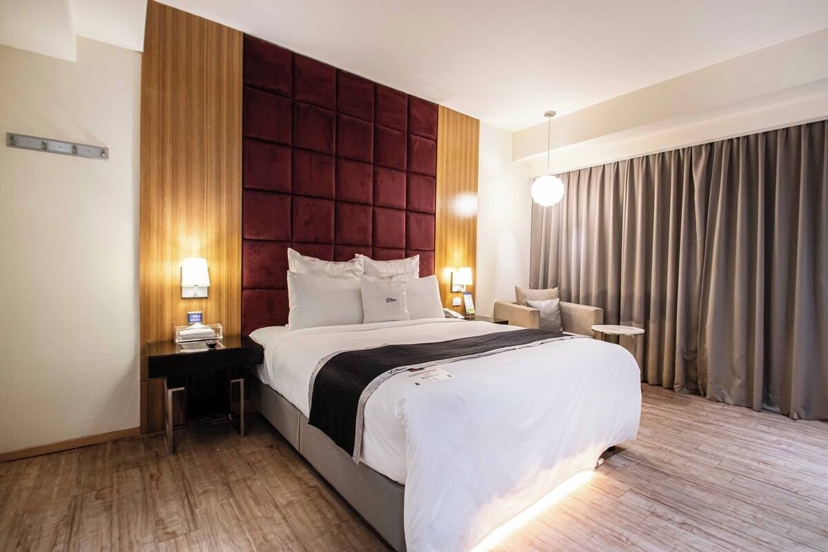 Hotelkamer met een groot bed, een houten hoofdbord, een bureau met lamp, een stoel en uitzicht buiten door gordijnen.