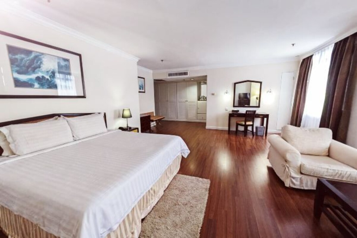 A hotel room with a large bed, a desk, a chair, and a sofa. There is a framed picture on the wall and a window with curtains.