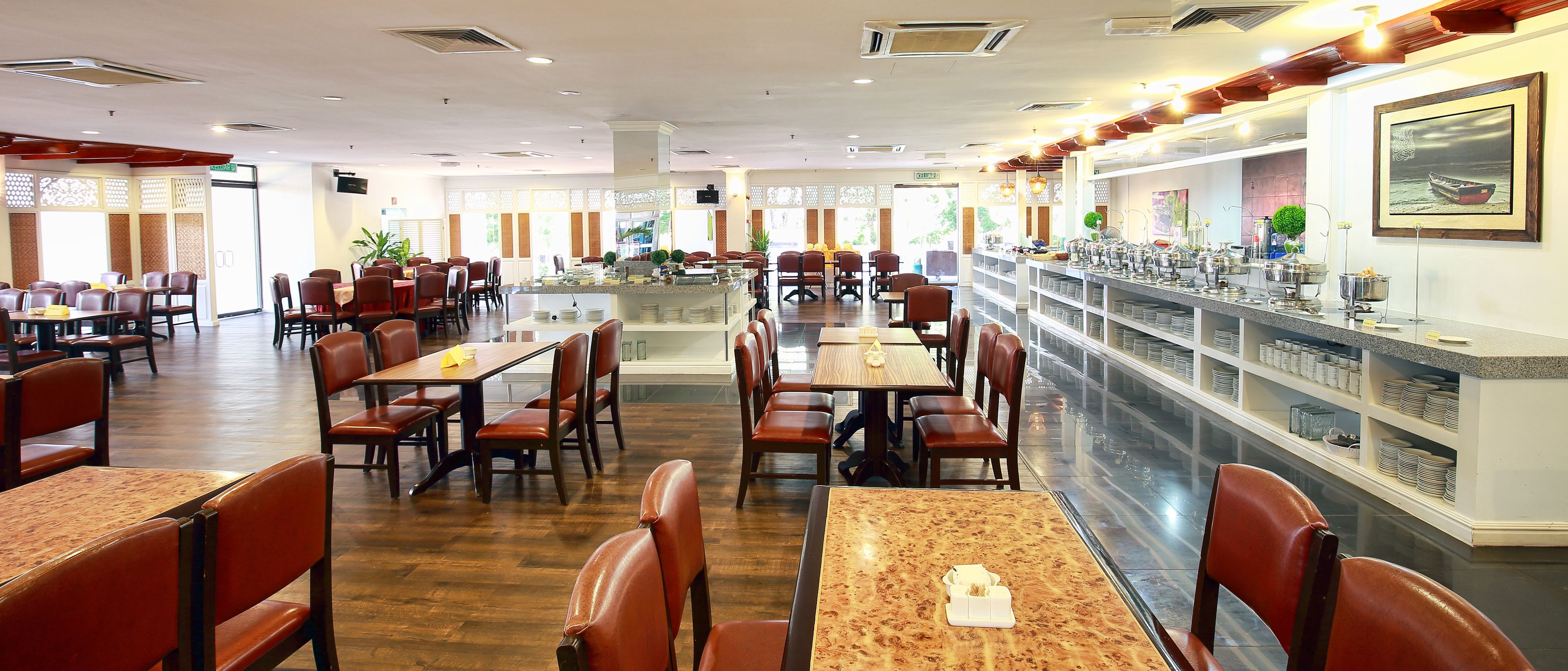 A spacious restaurant with multiple dining tables and chairs, a buffet counter with various dishes, and a large window providing natural light.