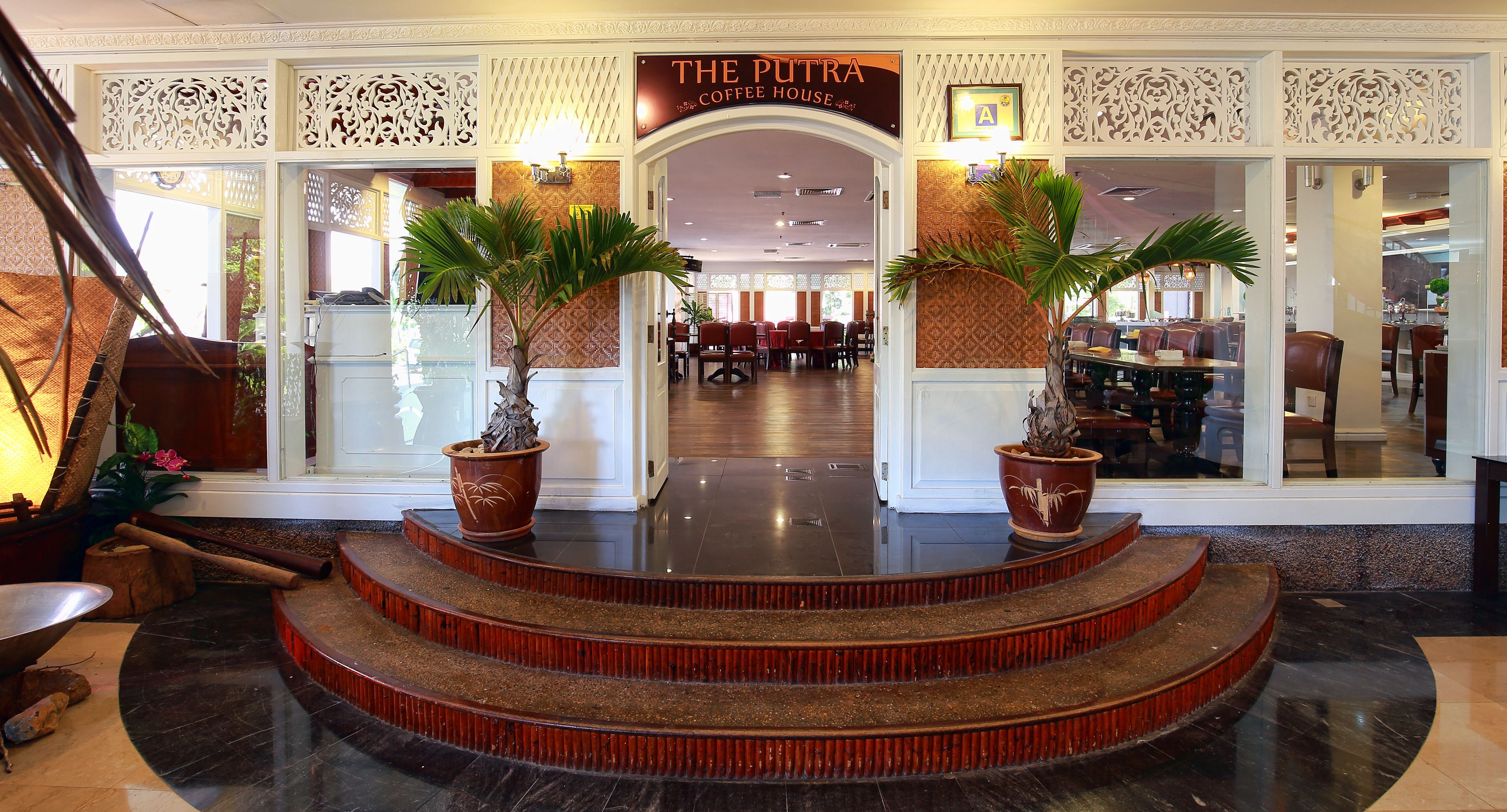 Entrada de un hotel con un letrero que dice "The Putra Coffee House", destacando una escalera con plantas en macetas y una pared decorativa.